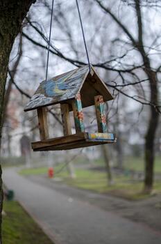 A rustic, painted wooden bird feeder hangs from a tree branch in an outdoor setting, with a blurred path and bare trees in the background. photo