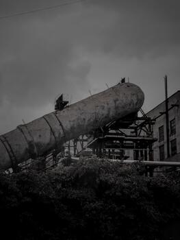 Dark, moody image of a deserted factory with overgrown vegetation and a large rusted pipe under a cloudy sky. photo