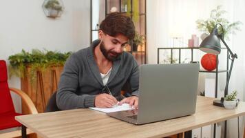 Indian man at home studies online with laptop copies notes from e-book to notebook focused calm. Arabian guy at desk writes by hand listens lesson keeps attention learns new skills progress video