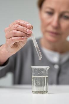 A middle-aged Caucasian woman adds a drop of reagent from a pipette into a beaker with a clear liquid. Laboratory surface, neutral background, educational experiment. photo