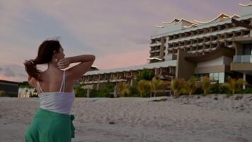 jong vrouw in wit halster top en groen broek poses met handen achter hoofd Aan zanderig strand Bij schemering, modern luxe toevlucht met gebogen architectuur achtergrond, vakantie levensstijl. video