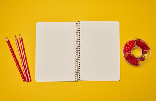 An open spiral-bound notebook with blank pages, surrounded by red pencils and paper clips on a yellow background. photo
