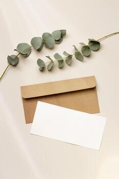 Blank paper card mock-up and craft envelope, eucalyptus plant on beige table. White empty letter or invitation, sunlight, beautiful soft shadow. Vertical lifestyle photo. Flat lay copyspace photo