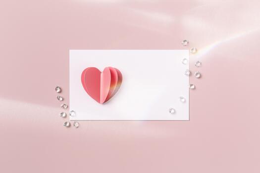 Pink heart and empty white paper card for love note with sequins, rhinestones on pink background with sunlight. Minimal style flat lay, mock up valentine card or wedding invitation. Romantic photo