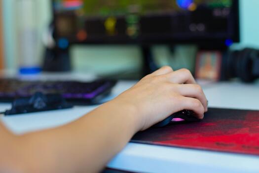 Close-up of a hand using a gaming mouse on a vibrant mouse pad, with a blurred computer monitor displaying a colorful gaming interface in the background, showcasing gaming culture photo