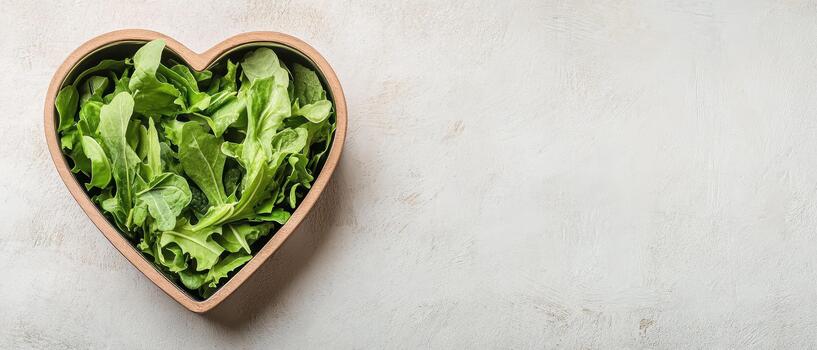 Arugula leaves fill heart shaped bowl. Fresh green salad in a wooden container on textured surface. Light background with copy space. photo