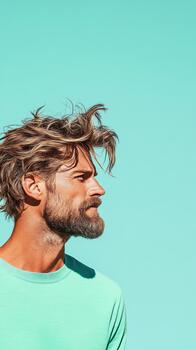 Man with messy hair and beard looks to the side on a bright turquoise background. photo