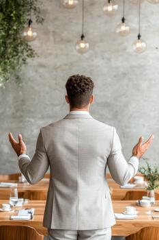 Man in light suit gesturing with open hands during a presentation in a modern room with hanging lights and plants. photo