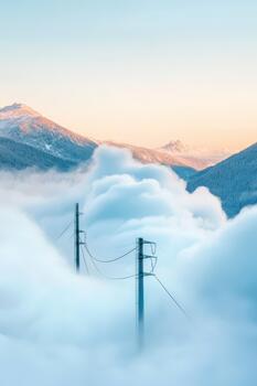 Two tall poles with wires stand amidst soft white clouds with snowy mountain peaks in the distance under a pale sky. photo