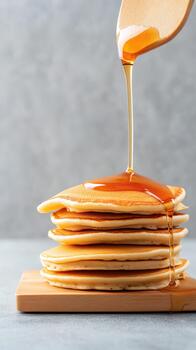 Stack of golden pancakes with sweet syrup pouring from a wooden spoon on a rustic board, grey textured background. photo
