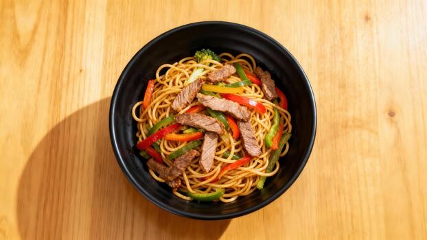 Overhead photograph of stir-fried lo mein noodles mixed with sliced beef, red and green bell peppers, and broccoli in a dark ceramic bowl placed on a wooden table. photo