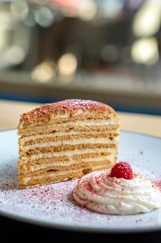 Layered honey cake slice topped with a raspberry served on a plate with cream. Elegant presentation against a blurred background. photo