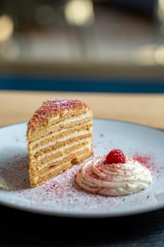 A slice of layered honey cake served on a plate with a swirl of cream and a raspberry garnish. photo