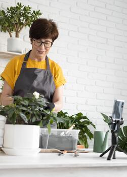 Middle aged woman blogger in front of smartphone camera on tripod records instructional tutorial for her blog shoots process of replanting flowers and plants full of soil enjoys botanic hobby photo