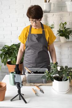 Female blogger standing in front of smartphone camera on tripod records instructional tutorial for her blog shoots process of replanting green plants full of soil enjoys botanic hobby photo