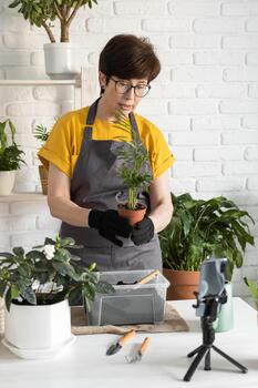 Woman blogger standing in front of smartphone camera on tripod records instructional tutorial for her blog shoots process of replanting flowers and green plants full of soil enjoys botanic hobby. photo
