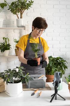 Middle aged woman blogger standing in front of smartphone camera on tripod records instructional tutorial for her blog shoots process of replanting flowers and plants of soil enjoys botanic hobby. photo