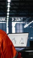 vertikal arbetstagare använder sig av automatisering programvara ansluten till robot maskineri via bärbar dator, omgiven förbi metall Utrustning och tillverkning verktyg. industriell miljö slingor teknik. kamera b. video
