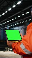 vertikal grön skärm läsplatta med arbetstagare hantera drift uppgifter tvärs över de fabrik golv, omgiven förbi metall maskineri och tillverkning verktyg den där markera professionell teknik. kamera a. video