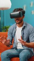 indisk man använder sig av virtuell verklighet trogen teknologi vr app headsetet hjälm skriver på virtuell tangentbord, spela simulering 3d 360 uppkopplad spel på Hem lägenhet. kille i glasögon Sammanträde på soffa video