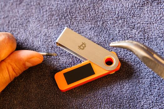 Light-skinned man focuses on tampering with a hardware wallet using a screwdriver and pliers. photo