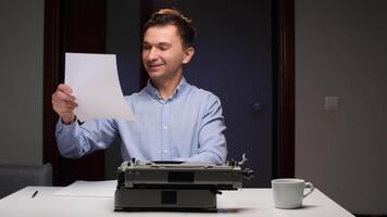 giovane uomo sorridente e lettura pagine, Tenere multiplo lenzuola di carta mentre seduta a un' scrivania con un vecchio macchina da scrivere e un' caffè tazza, impegnato nel creativo scrittura opera video