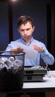 vertical 1 homem desintegra-se uma peça do papel, expressando frustração enquanto sentado às uma escrivaninha com a velho máquina de escrever e uma cheio cesto de lixo, experimentando escritoras quadra video