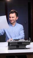 vertikal leende man granskning en stack av papper medan Sammanträde på en skrivbord med en retro mekanisk skrivmaskin, njuter de bearbeta av skrivning och skapande video