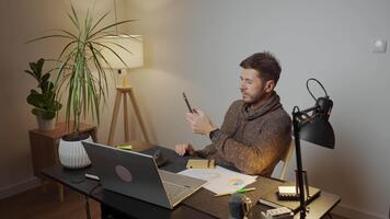 man Sammanträde på skrivbord använder sig av smartphone medan arbetssätt i en Hem kontor med bärbar dator och dokument. trött frilansare förhalar på arbetsplats under avlägsen arbete. improduktiv man använder sig av telefon video