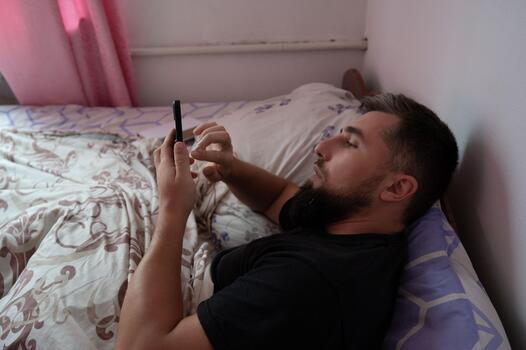 Man in bed using his phone with a focused expression. The scene is in a well-lit room with floral bedding adding a touch of comfort. photo
