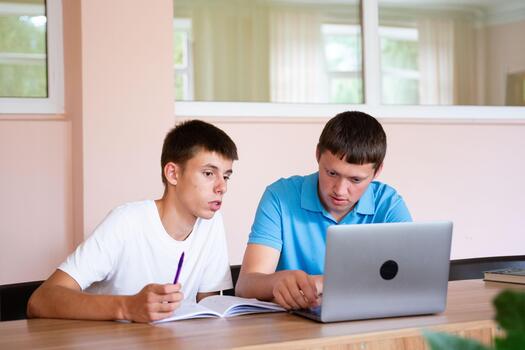 Two young men collaborate on a laptop at a desk, one with pen and notebook. Study partners working on a project together, indoors. photo