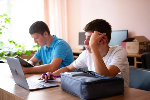 Teenagers studying at a table with a laptop and books. One student looks pensive while the other focuses on reading during their study session. photo
