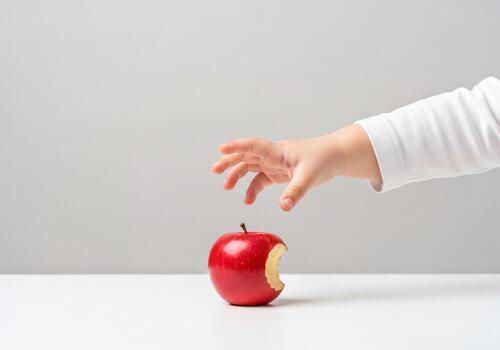 Child's small hand reaching towards a fresh red apple with a bite mark on white photo
