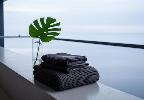 Stack of Dark Grey Waffle Pattern Towels and Monstera Leaf in Serene Modern Spa Setting with Water View photo