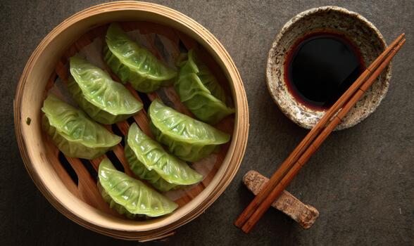 A bowl of green dumplings sits on a table with chopsticks and a dipping sauce photo