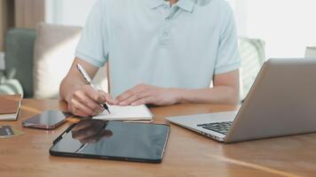 een Mens is schrijven Aan een notitieboekje terwijl zittend Bij een tafel met een laptop en een pen video