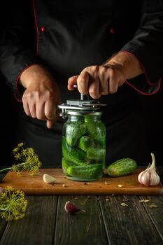 A man uses a manual seaming key to seal a jar of pickled cucumbers on a kitchen counter. Concept of pickling cucumber with garlic and dill photo