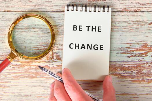 A hand holds a pencil next to an open notepad displaying the powerful phrase Be the change. A magnifying glass rests nearby, symbolizing focus on growth and action in life photo