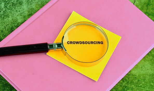 A magnifying glass focuses on the word crowdsourcing written on a yellow sticky note placed atop a pink notebook. This setup creates a visually engaging display photo