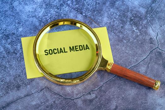 A magnifying glass is placed over a yellow sheet with the text social media printed in bold. The background features a textured surface that adds depth photo