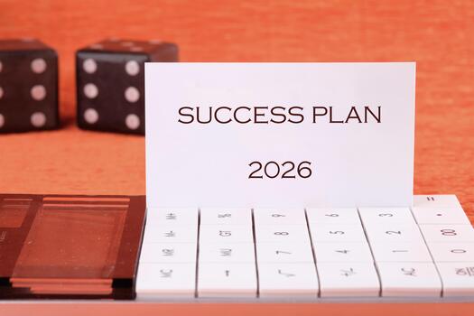 A strategic guide is displayed, titled Success Plan 2026, resting on a sleek calculator. Nearby, two dice add a playful element, suggesting that success involves chance and planning photo