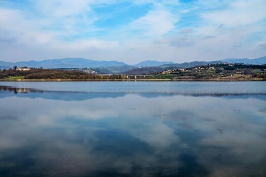 A serene mountain lake reflecting a bright blue sky and distant hills in a quiet rural setting. The calm water creates a mirror-like surface, photo