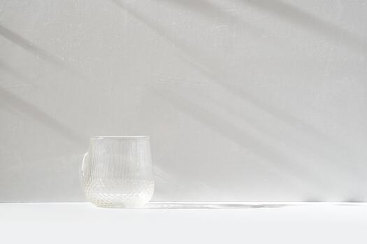Minimalist still life of an empty textured glass mug on a white surface with long soft shadows on a light grey wall, creating a calm and airy atmosphere with natural daylight photo