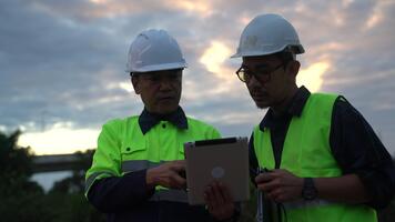 Two construction engineers or supervisors in high-visibility vests collaboratively reviewing digital plans and schematics on a tablet computer at the job site during twilight, emphasizing teamwork video