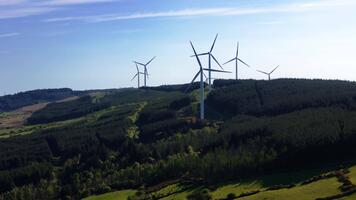 Wind turbines stand atop a tree covered hill on a sunny day in Ireland. The turbines are turning, generating power. Blue sky with light clouds is visible. video