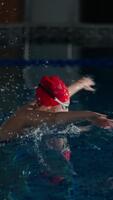 Focused swimmer in red cap gliding through dark indoor pool water, creating powerful splashes and ripples while practicing intense lap swimming technique video