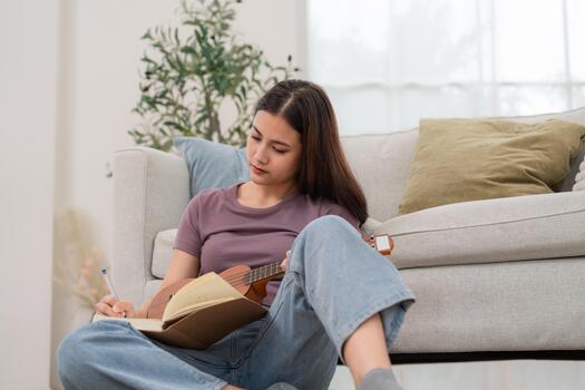 Young person writes in a notebook while playing a ukulele at home during the afternoon photo
