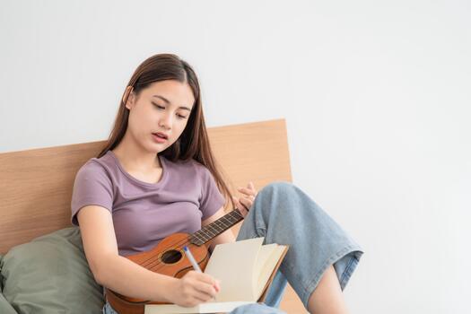 Young woman plays ukulele and writes in notebook while sitting on bed in simple room during daytime hours photo