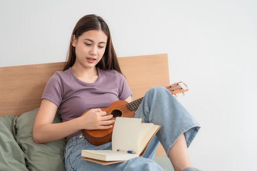 Young woman plays ukulele and writes in notebook while sitting on a bed in a simple indoor setting photo