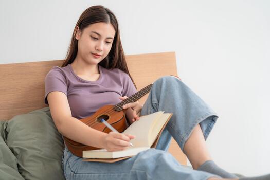 Young person writes in notebook while sitting on bed and playing ukulele in a simple indoor setting photo